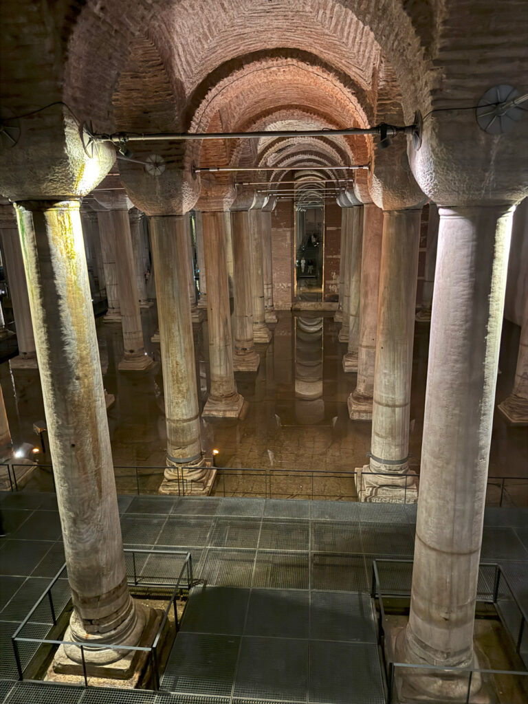 A view of the Basilica Cistern entrance hall in Istanbul, featuring tall marble columns reflected in the water under arched brick ceilings.