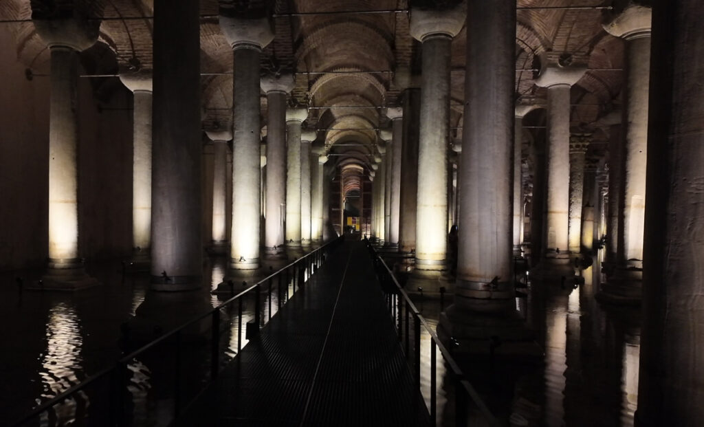 A dramatic view of the Imperial Walkway inside the Basilica Cistern in Istanbul, with towering marble columns illuminated by soft lights reflecting on the water.