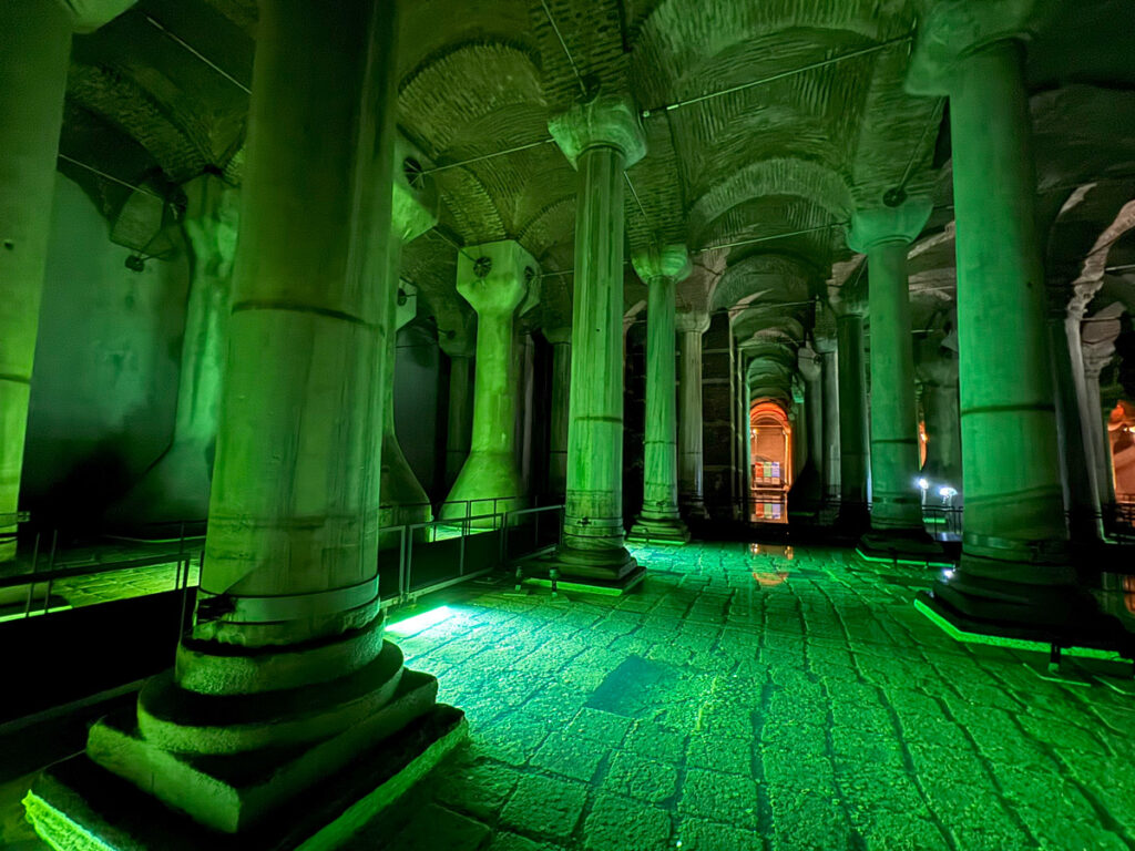 Illuminated waterways inside the Basilica Cistern, where green lights reflect off the stone floor and towering columns, creating a surreal mirror-like effect.