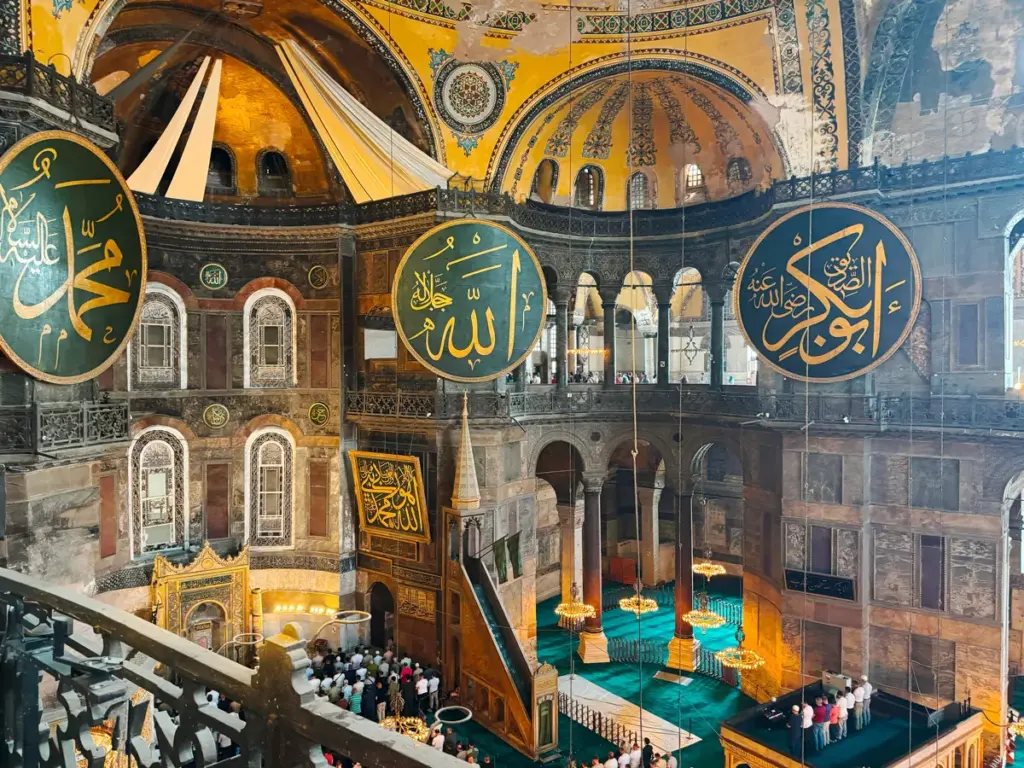 Interior of the Hagia Sophia in Istanbul, showing the grand dome, Islamic calligraphy medallions, and sunlight filtering through the upper windows.
