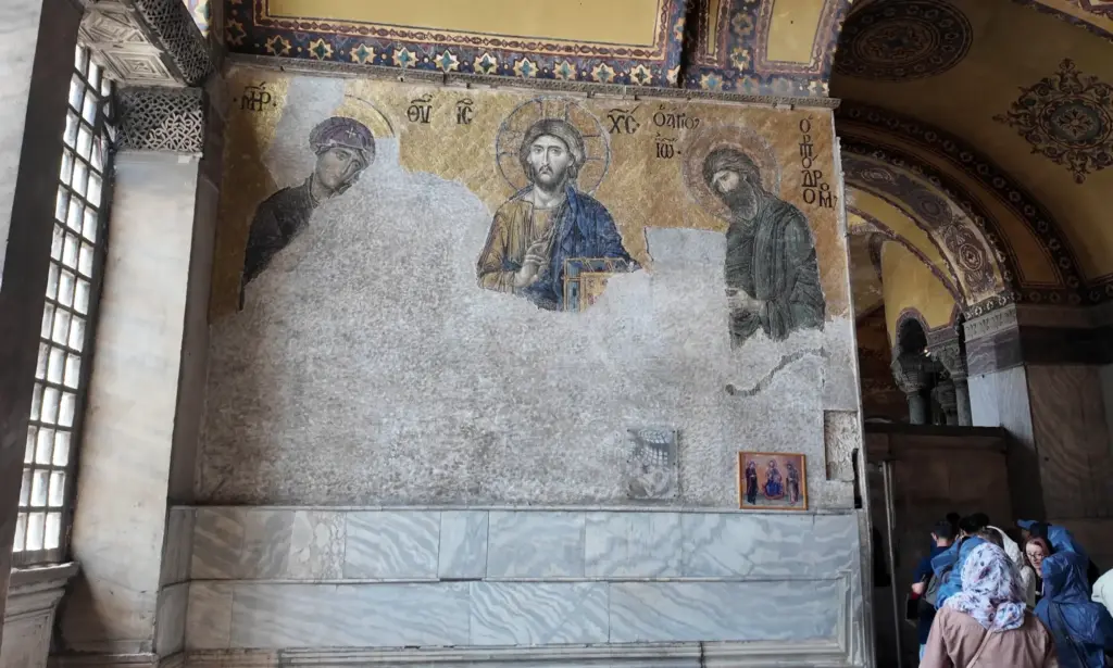 The Deësis mosaic in the upper south gallery of Hagia Sophia, showing Christ, the Virgin Mary, and John the Baptist, partially damaged but still radiant under natural light.